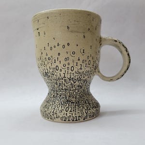 May include: A beige ceramic mug with an hourglass shape. The mug features a pattern of black binary code numbers, creating a modern design. The handle has a textured finish. The mug is suitable for hot drinks.