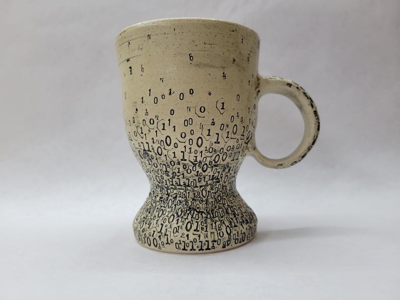 May include: A beige ceramic mug with an hourglass shape. The mug features a pattern of black binary code numbers, creating a modern design. The handle has a textured finish. The mug is suitable for hot drinks.