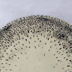 May include: A ceramic plate with a cream-colored surface and a black binary code pattern around the rim. The numbers 0 and 1 are arranged in a repeating pattern, creating a unique design. The plate's design is a modern take on digital art.