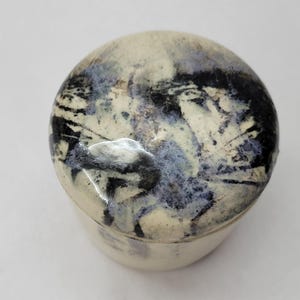 Könnte beinhalten: Ein kleines, rundes Keramikgefäß mit Deckel und glänzender Oberfläche. Das Design ist abstrakt und malerisch in Creme-, Tiefschwarz- und gedämpften Violetttönen gehalten, mit dunklen, streifigen Pinselstrichmustern auf dem Deckel.