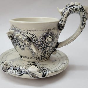May include: A ceramic cup and saucer set in off-white with intricate, raised dark grey and black textured detailing. The cup features a sculpted handle and a complex, relief-style pattern covering the exterior surface and the saucer rim.