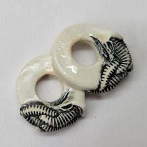 May include: Two circular, off-white ceramic beads featuring a detailed, raised black relief design of an elephant trunk and tusk wrapping around one side of each ring. The surface of the ceramic has a smooth, glossy finish with dark accents.