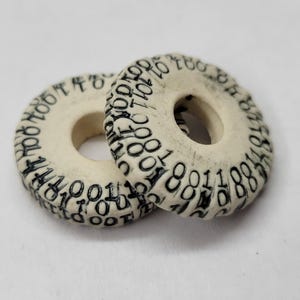 Puede incluir: Dos cuentas circulares de color blanquecino con un agujero en el centro. Cada cuenta está cubierta de números negros alrededor del borde exterior. Las cuentas probablemente se utilizan para la fabricación de joyas o manualidades.