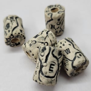 May include: A collection of off-white cylindrical beads with black markings. The beads feature a map-like design with directional letters like N, E, S, and W. Each bead has a hole running through the center, suitable for stringing.