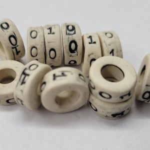 Puede incluir: Cuentas en forma de anillo de color marfil con números negros. Las cuentas están dispersas, mostrando números como 0, 1 y 9. Estas cuentas podrían usarse para hacer joyas u otras manualidades.