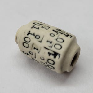 May include: A cylindrical off-white bead featuring a repeating black binary code pattern of ones and zeros printed around its exterior surface. The bead has a central hole running through its length and a slightly recessed design at both ends.