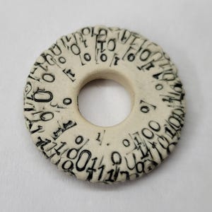 May include: A cream-colored, circular pendant with a central hole. The pendant is adorned with a repeating pattern of black numbers and symbols around its circumference. The design gives the pendant a unique, textured appearance.