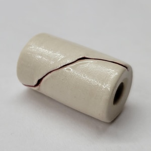 May include: A cylindrical, off-white ceramic bead with a dark red crack running diagonally across its surface. The bead has a hole through its center and a glossy finish. The crack adds a unique, artistic element to the bead.