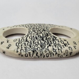 May include: An oval-shaped ceramic button with a smooth off-white finish. The surface is decorated with a dense, repeating black print of binary code consisting of zeros and ones. The button features two large, circular openings in the center.