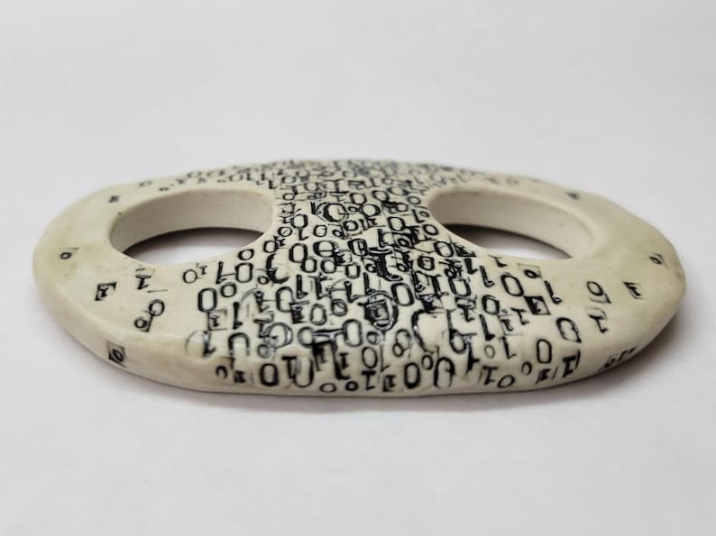 May include: An oval-shaped ceramic button with a smooth off-white finish. The surface is decorated with a dense, repeating black print of binary code consisting of zeros and ones. The button features two large, circular openings in the center.