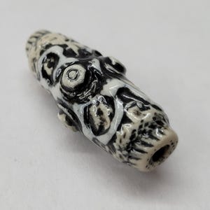 May include: A cylindrical ceramic bead featuring a high-contrast black and off-white marbled pattern. The surface has a glossy finish with raised, textured details and a central hole running through the length of the bead for threading.