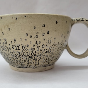 May include: A beige ceramic coffee mug with a large handle. The mug is decorated with black binary code numbers, with the numbers densely packed near the bottom and sparse near the top. The mug has a wide, rounded shape.