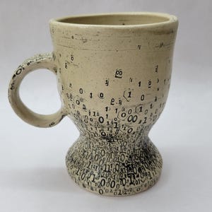 May include: A handcrafted ceramic mug with a beige glaze and a distinctive design. The mug has a handle and a flared base, decorated with a pattern of black numbers and symbols. The numbers resemble binary code, giving it a tech-inspired look.