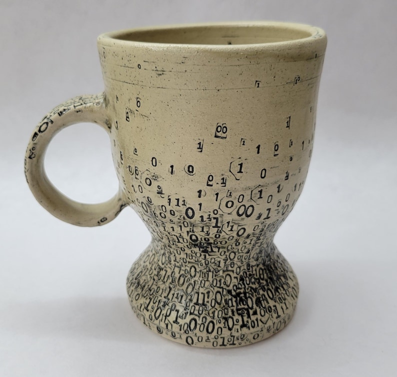 May include: A handcrafted ceramic mug with a beige glaze and a distinctive design. The mug has a handle and a flared base, decorated with a pattern of black numbers and symbols. The numbers resemble binary code, giving it a tech-inspired look.