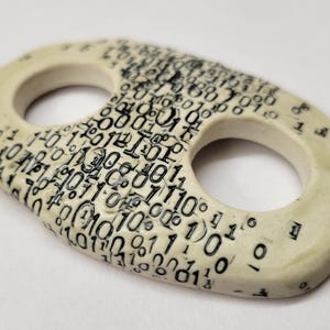 Peut inclure: Un bouton en céramique de forme ovale avec deux trous circulaires, présentant une couleur de base blanc cassé et un motif dense imprimé en noir de code binaire composé de zéros et de uns couvrant toute la surface supérieure de l'objet.