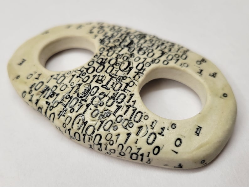 May include: An oval-shaped ceramic button with two circular holes, featuring an off-white base color and a dense, black printed pattern of binary code consisting of zeros and ones covering the entire top surface of the object.