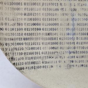 May include: A curved, off-white ceramic plate featuring a repeating pattern of black binary code consisting of zeros and ones printed across the entire surface. The design has a slightly distressed, textured appearance with visible ink variations.