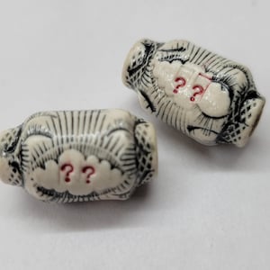 May include: Two off-white ceramic beads with black etched designs and red question marks. The beads have a cylindrical shape with rounded ends and are decorated with a cloud-like design.