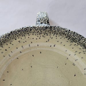 May include: A ceramic bowl with a cream-colored interior and a textured rim featuring a dense, black-printed pattern of binary code digits zero and one. The design transitions from a concentrated, layered border to sparse, scattered digits toward the center.
