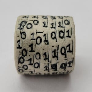 Puede incluir: Un objeto cilíndrico con una superficie beige claro, grabado con números de código binario negro. Los números están dispuestos en columnas verticales alrededor del cilindro. El objeto parece ser un artículo único, hecho a mano.