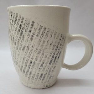 May include: Cream-coloured ceramic mug with a rounded handle. The mug features a pattern of black binary code. The design covers most of the mug's surface, creating a tech-inspired aesthetic. The mug is suitable for hot drinks.