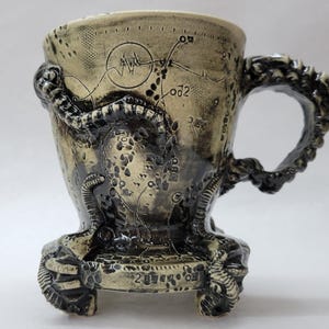 May include: A beige ceramic mug with a dark, textured glaze featuring intricate black line drawings of maps, symbols, and a heartbeat monitor. The mug has a sculpted handle and base resembling organic, tentacle-like forms with dark detailing.