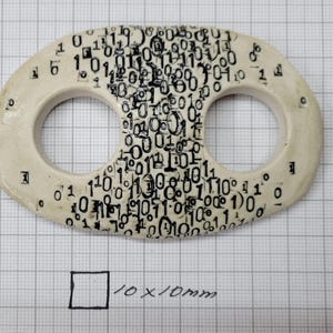 May include: An oval-shaped off-white ceramic toggle with two circular openings, featuring a dense black printed pattern of binary code digits zero and one. A hand-drawn square labeled 10x10mm is visible below the toggle on the grid paper.