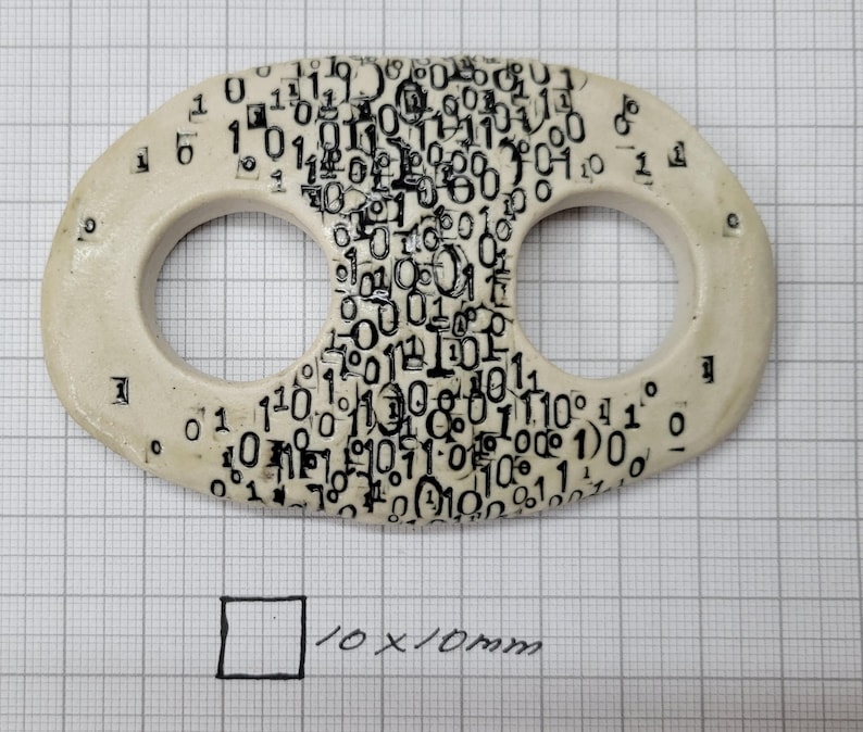 May include: An oval-shaped off-white ceramic toggle with two circular openings, featuring a dense black printed pattern of binary code digits zero and one. A hand-drawn square labeled 10x10mm is visible below the toggle on the grid paper.