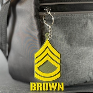 May include: A yellow and green keychain featuring a military rank insignia and the name "BROWN" in yellow block letters. The keychain is attached to a silver chain and a silver keyring. The background is a gray bag.