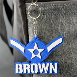 May include: A blue and white keychain with the name "BROWN" in white block letters. The keychain features a star and wing design, attached to a silver chain and keyring. The keychain is hanging against a gray background.