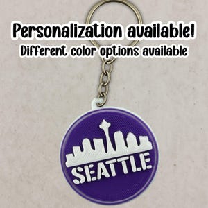May include: A purple and white Seattle keychain with a silver keyring. The keychain features the Seattle skyline and the word "SEATTLE" in white. Text on the image reads "Personalization available! Different color options available."