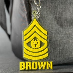 May include: A yellow and green keychain featuring a military rank insignia with the name "BROWN" in yellow block letters. The keychain is attached to a silver keyring and is set against a gray background.