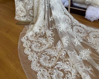 May include: White lace fabric with intricate floral and scrollwork embroidery. The delicate, sheer material is draped over a mannequin, showcasing the detailed design. Rolls of white fabric are visible in the background, suggesting a bridal or formal wear theme.