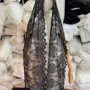 Peut inclure: Un voile en dentelle noire avec un bord festonn&eacute; est drap&eacute; sur un mannequin. Le motif floral complexe de la dentelle est visible. L'arri&egrave;re-plan montre des piles de tissu blanc.