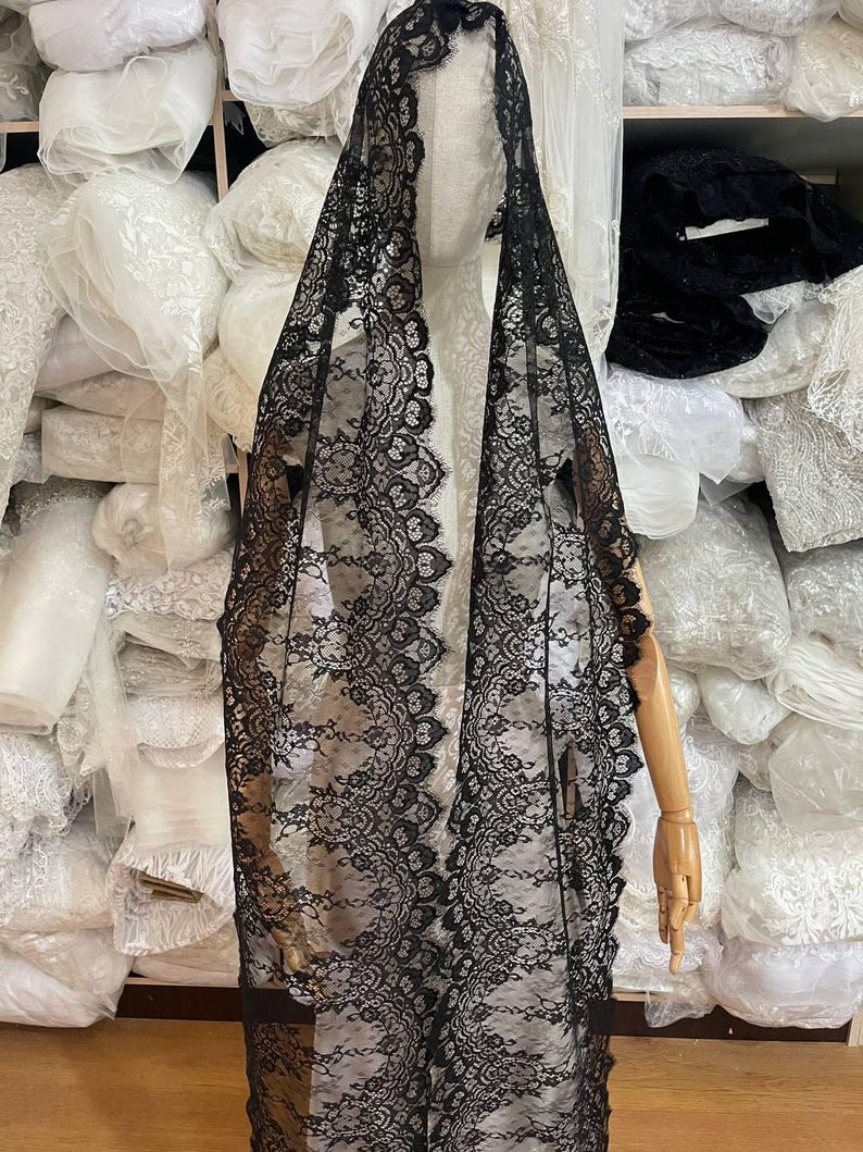 Peut inclure: Un voile en dentelle noire avec un bord festonn&eacute; est drap&eacute; sur un mannequin. Le motif floral complexe de la dentelle est visible. L'arri&egrave;re-plan montre des piles de tissu blanc.