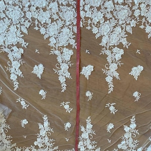 Puede incluir: Tela de encaje floral marfil con un dise&ntilde;o delicado y tridimensional. El encaje presenta patrones detallados de flores y hojas blancas. Una cinta m&eacute;trica roja se coloca verticalmente, indicando la longitud de la tela.
