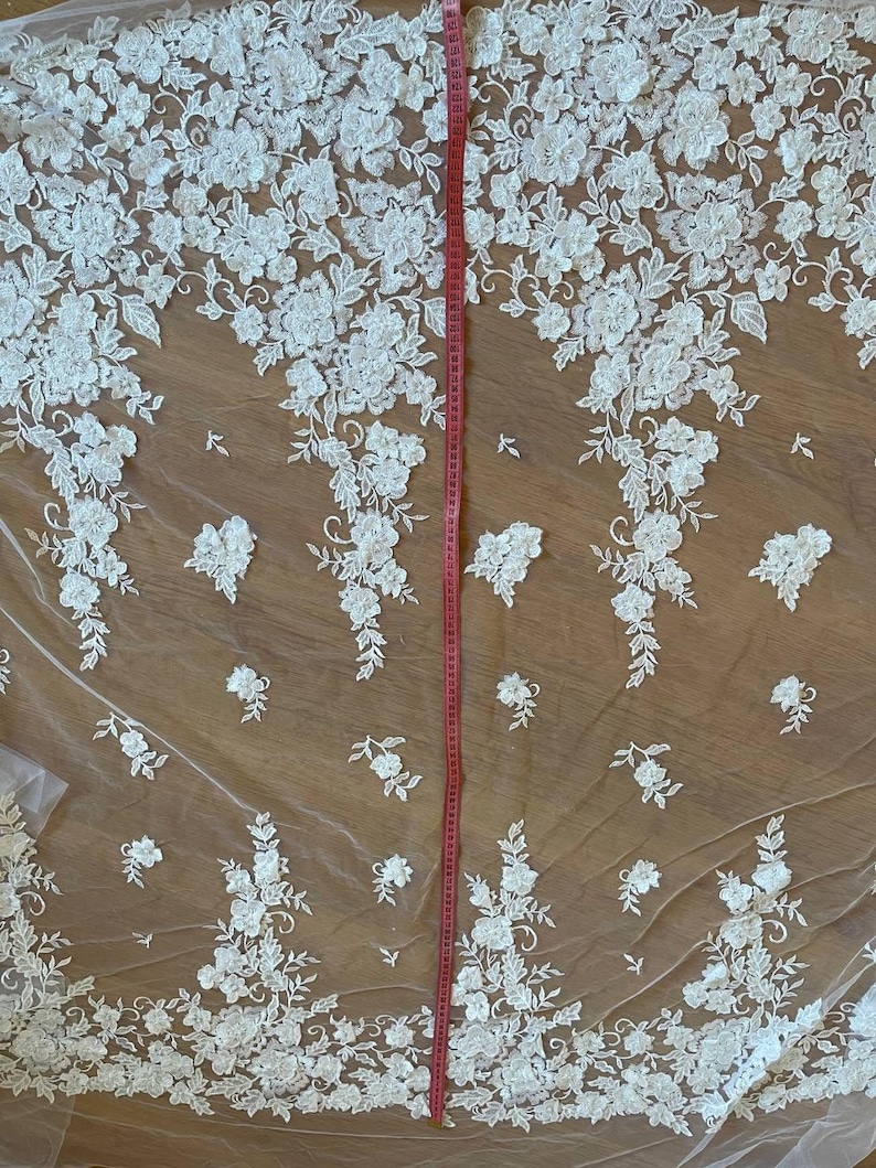 Puede incluir: Tela de encaje floral marfil con un dise&ntilde;o delicado y tridimensional. El encaje presenta patrones detallados de flores y hojas blancas. Una cinta m&eacute;trica roja se coloca verticalmente, indicando la longitud de la tela.