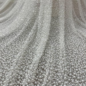 May include: Close-up of white fabric densely covered in small, round, white beads. The beads vary in size, creating a textured surface. The fabric is draped, showing folds and shadows, and is suitable for bridal or formal wear.