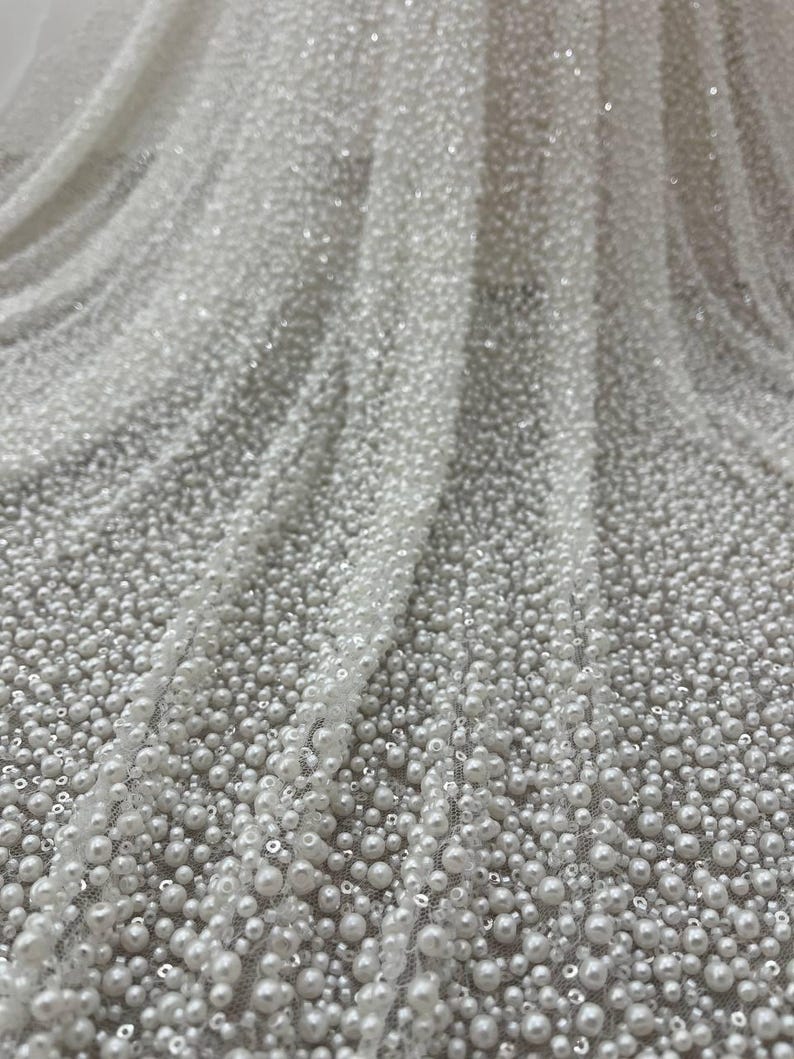 May include: Close-up of white fabric densely covered in small, round, white beads. The beads vary in size, creating a textured surface. The fabric is draped, showing folds and shadows, and is suitable for bridal or formal wear.