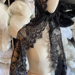 Peut inclure: Un ch&acirc;le en dentelle noire drap&eacute; sur un mannequin. Le ch&acirc;le pr&eacute;sente un motif floral d&eacute;licat sur les bords et est fait d'un tissu l&eacute;ger et transparent. Des rouleaux de tissu blanc sont visibles en arri&egrave;re-plan.