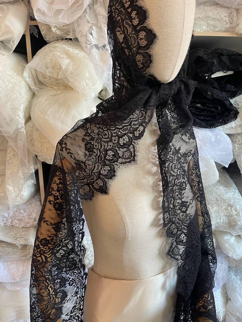 Peut inclure: Un ch&acirc;le en dentelle noire drap&eacute; sur un mannequin. Le ch&acirc;le pr&eacute;sente un motif floral d&eacute;licat sur les bords et est fait d'un tissu l&eacute;ger et transparent. Des rouleaux de tissu blanc sont visibles en arri&egrave;re-plan.
