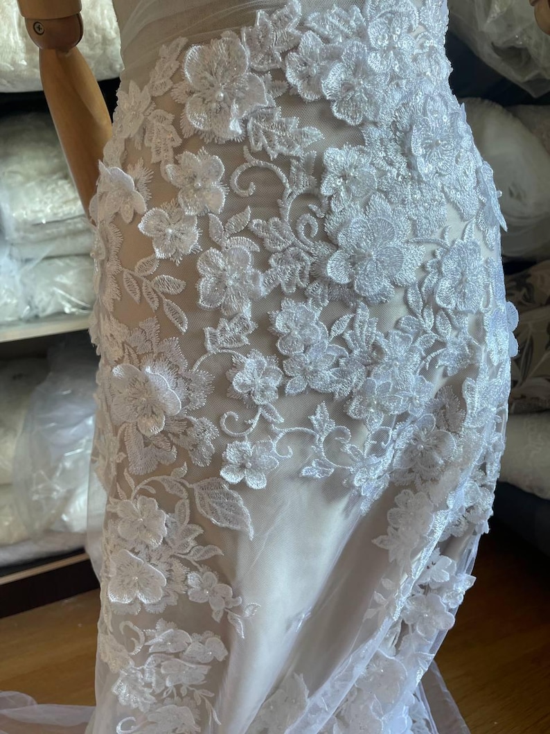 3D floral lace fabric with embroidered flowers and delicate beaded details, white lace material by the yard for sewing wedding dresses and bridal gowns, luxury lace with 3D floral embroidery for creating elegant and romantic dress designs, soft and high quality fabric perfect for veils, skirts, overlays and evening gowns, bridal lace fabric with sequins and beads for couture sewing projects, wedding lace with floral texture that adds volume and sophistication to any gown
