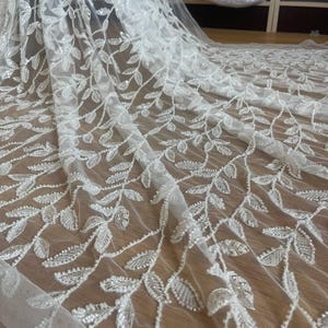 Könnte beinhalten: Weißer Tüllstoff mit einem gestickten Blattmuster. Die Blätter sind mit kleinen, glitzernden Perlen verziert, die eine zarte und elegante Textur erzeugen. Der Stoff ist drapiert und zeigt seinen Fall, ideal für Braut- oder Abendmode.