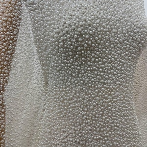 May include: Close-up of a white fabric densely covered in small, round, pearl-like beads. The beads vary slightly in size, creating a textured surface. The fabric appears to be draped, suggesting a garment or decorative textile.