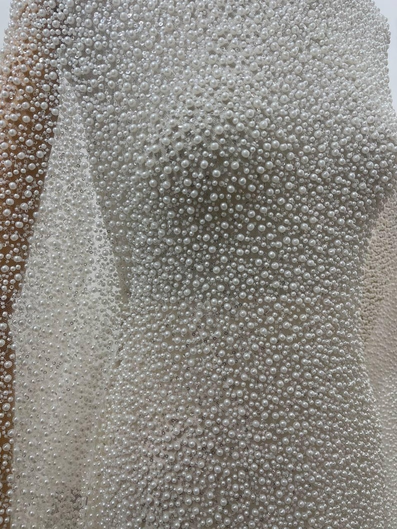 May include: Close-up of a white fabric densely covered in small, round, pearl-like beads. The beads vary slightly in size, creating a textured surface. The fabric appears to be draped, suggesting a garment or decorative textile.