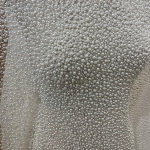 May include: Close-up of a white fabric densely covered in small, round, pearl-like beads. The beads vary slightly in size, creating a textured surface. The fabric appears to be a bridal or formal wear material.