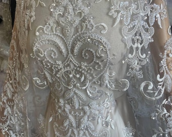 May include: Close-up of white lace fabric with intricate floral and scrollwork embroidery. The lace is embellished with sequins and beads, creating a shimmering effect. The fabric is draped over a dress form, showcasing its delicate texture and design.