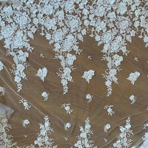 Puede incluir: Tela de encaje floral blanco con un delicado patr&oacute;n de flores y hojas. El encaje est&aacute; dispuesto en un patr&oacute;n repetitivo, con las flores agrupadas en grupos. La tela es semitransparente y el fondo es de color marr&oacute;n claro.