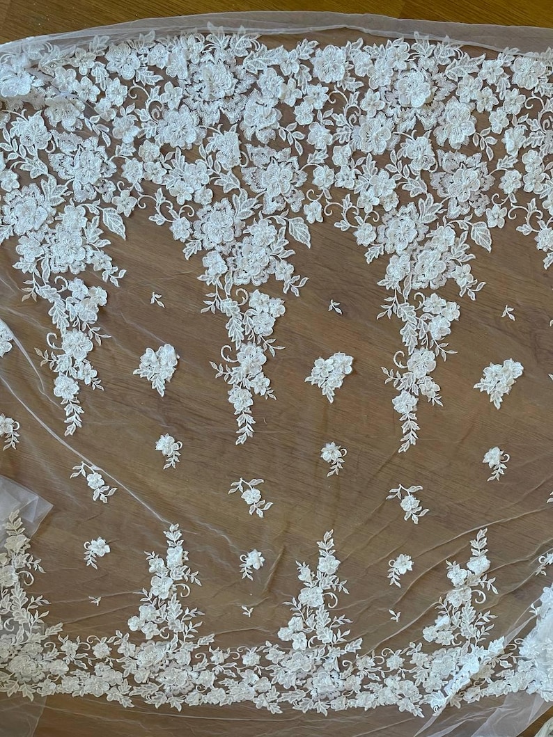 Puede incluir: Tela de encaje floral blanco con un delicado patr&oacute;n de flores y hojas. El encaje est&aacute; dispuesto en un patr&oacute;n repetitivo, con las flores agrupadas en grupos. La tela es semitransparente y el fondo es de color marr&oacute;n claro.
