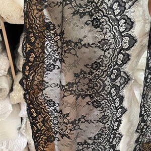 Peut inclure: Gros plan sur un tissu en dentelle noire avec un motif floral. La dentelle a un bord festonn&eacute; et est superpos&eacute;e &agrave; un tissu blanc. Le design complexe et la texture d&eacute;licate sont visibles. Le tissu est probablement utilis&eacute; pour l'habillement ou la d&eacute;coration int&eacute;rieure.